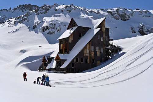 A group of people near a weathered wooden building in a snowy landscape, surrounded by mountains under a clear blue sky.