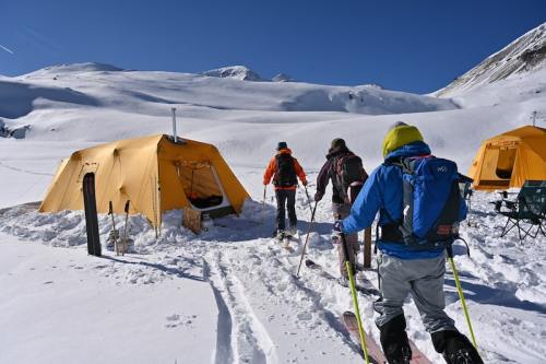 Three skiers walk towards yellow tents in a snowy landscape under a clear blue sky.