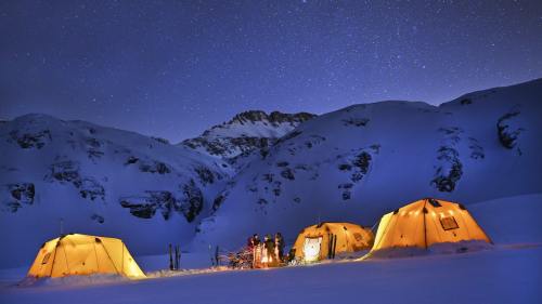 Tents illuminated under a starry night sky, with people gathered around a fire in a snowy mountain landscape.