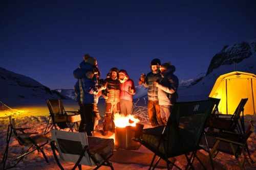 A group of people gathered around a campfire at night, with a tent and snowy landscape in the background.