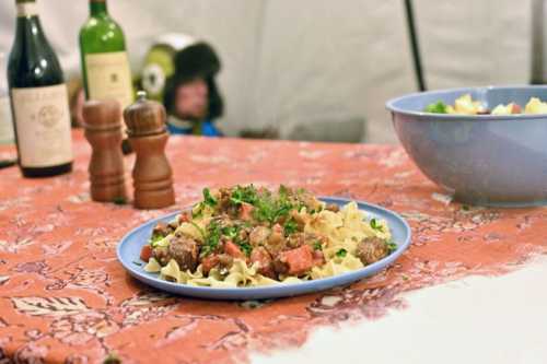 A plate of pasta with meat and vegetables, garnished with parsley, on a table with wine bottles and a salad in the background.