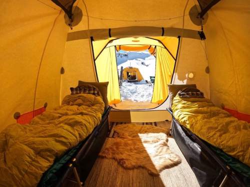 Cozy tent interior with two beds, warm blankets, and a view of snowy outdoors through the entrance.