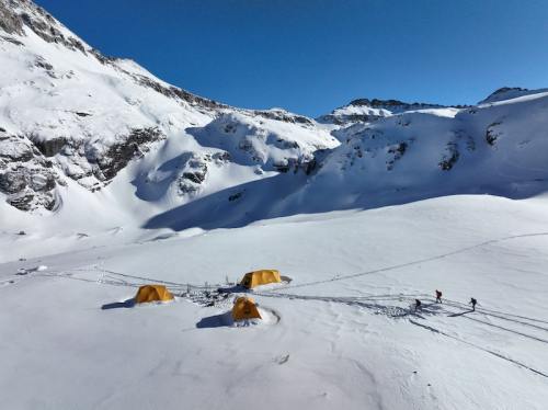 A snowy mountain landscape with yellow tents and two figures walking on a snow-covered terrain under a clear blue sky.