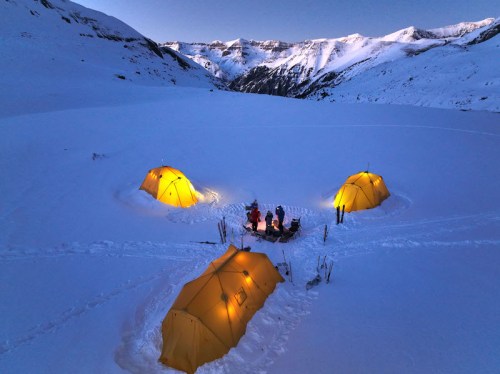 Three yellow tents illuminated at dusk, surrounded by snow-covered mountains and a small gathering of people.