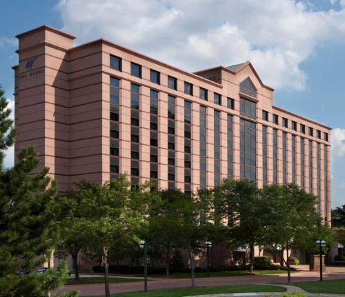A modern hotel building with large windows, surrounded by trees and a blue sky with clouds.