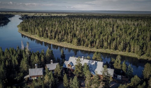 Aerial view of a river winding through a dense forest, with a rustic cabin nestled among the trees.