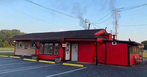 A red and wooden building with smoke rising from a chimney, surrounded by a parking lot and planters.