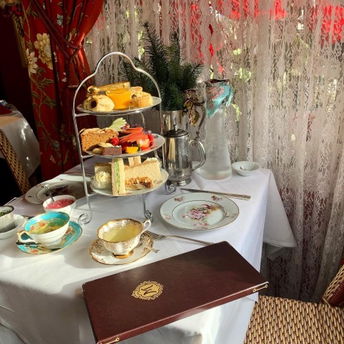 A beautifully arranged afternoon tea set with tiered treats, fine china, and a menu on a table by a lace curtain.