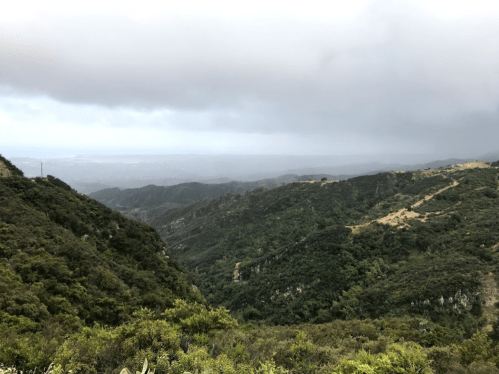 A scenic view of rolling green hills and valleys under a cloudy sky, with distant mountains in the background.