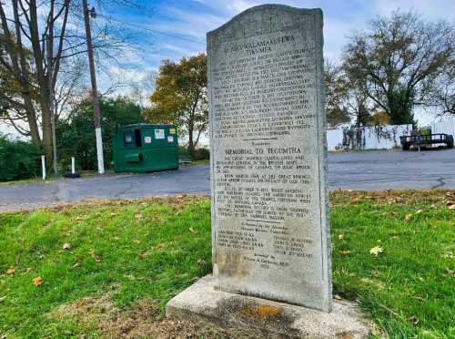 A stone memorial with inscriptions, set in a grassy area near a dumpster and trees under a cloudy sky.