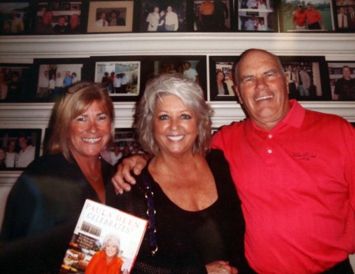 Three smiling individuals pose together in front of a wall of framed photos, holding a cookbook.