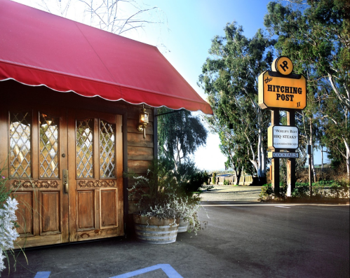 A rustic restaurant entrance with a red awning and a sign for "The Hitching Post II" in a sunny outdoor setting.