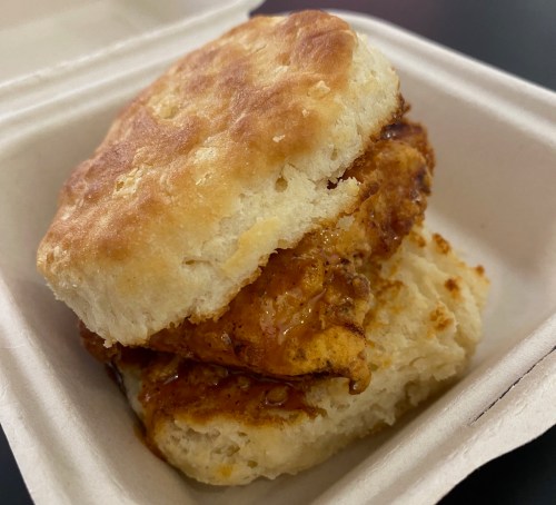 A close-up of a biscuit sandwich filled with crispy fried chicken and a spicy sauce, served in a takeout container.