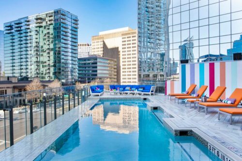 Rooftop pool with orange lounge chairs, surrounded by modern buildings and a clear blue sky.