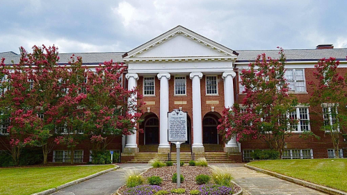 Historic brick building with white columns, surrounded by flowering trees and a grassy area.