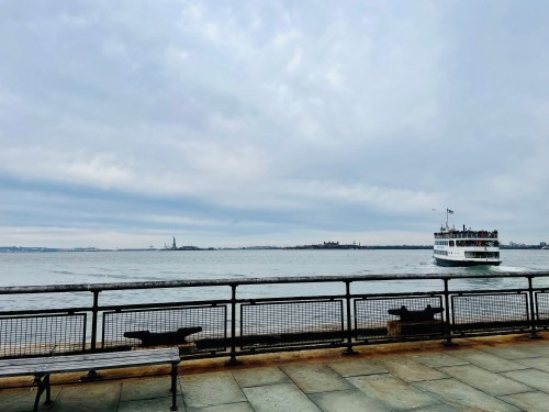 A ferry sails on a calm river with a cloudy sky, featuring benches along the waterfront. The Statue of Liberty is visible in the distance.