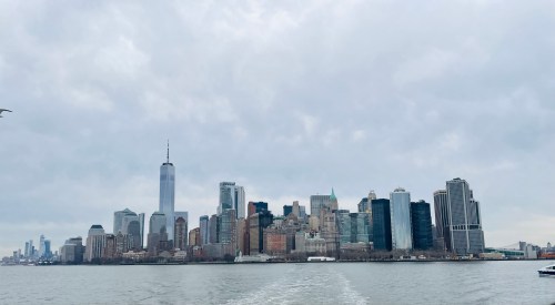 A panoramic view of the New York City skyline over a calm river, with cloudy skies and tall buildings.