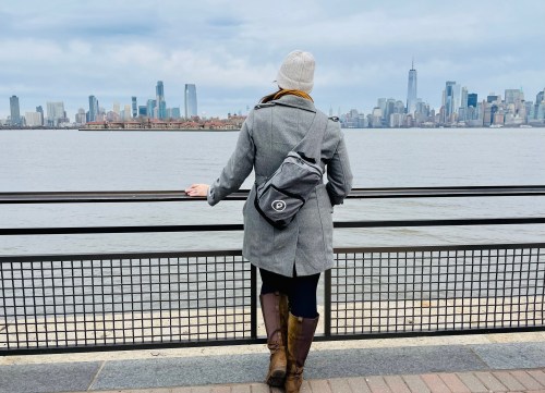 A person in a gray coat and beanie stands by a waterfront, gazing at a city skyline across the water.