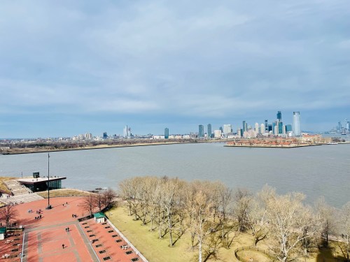 A view of a river with a city skyline in the background, featuring bare trees and a park area in the foreground.