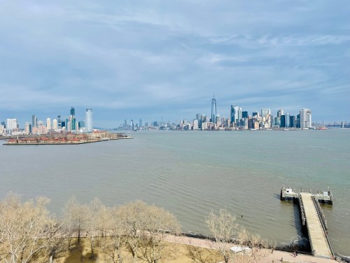 A view of the New York City skyline across the water, with a pier in the foreground and cloudy skies above.