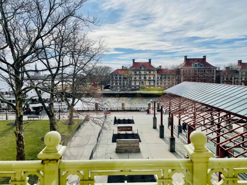 View of a waterfront area with historic buildings, trees, and a pavilion under a partly cloudy sky.