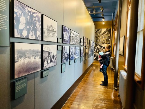A person holding a child stands in a museum, observing historical photographs displayed on the wall.
