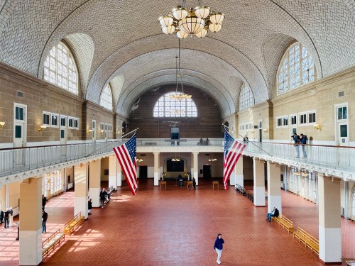 A spacious historic hall with high ceilings, large windows, and American flags, featuring visitors and benches.