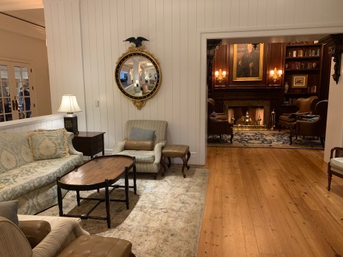 Cozy living room with a vintage sofa, armchairs, a round table, and a mirror, leading to a warmly lit library.
