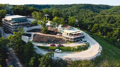 Aerial view of a hillside resort with multiple buildings, surrounded by greenery and a winding road.