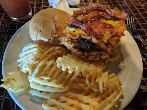 A plate with a fried chicken sandwich topped with bacon and cheese, served with waffle fries and a drink in the background.