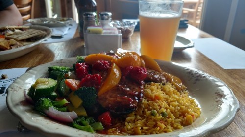 A plate of colorful stir-fried vegetables, grilled chicken with sauce, and yellow rice, alongside a glass of beer.