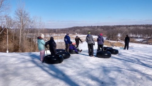 A group of people stands on a snowy hill with inner tubes, preparing to go tubing down the slope on a sunny day.
