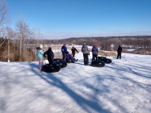 A group of people on a snowy hill preparing to go tubing, with trees and a landscape in the background.