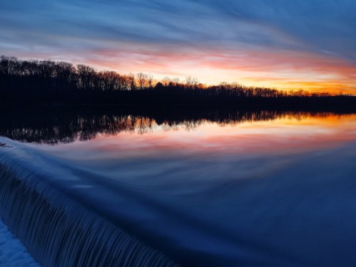 A serene sunset over a calm lake, with vibrant colors reflecting on the water and a gentle waterfall in the foreground.