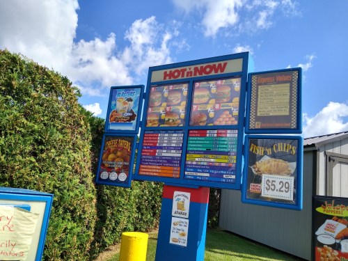 Menu board at a fast-food restaurant featuring burgers, fish and chips, and various combo meals against a blue sky.