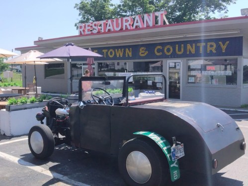 A vintage car parked in front of a restaurant with a sign reading "Town & Country." Outdoor seating is visible.