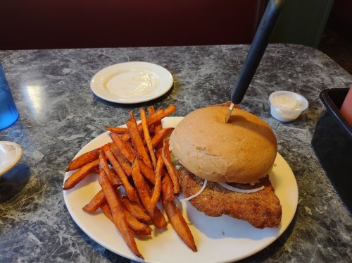 A plate with a fried fish sandwich topped with onions and a side of sweet potato fries.