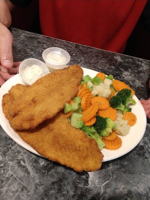 A plate with two fried fish fillets, a side of mixed vegetables, and two small cups of dipping sauce.