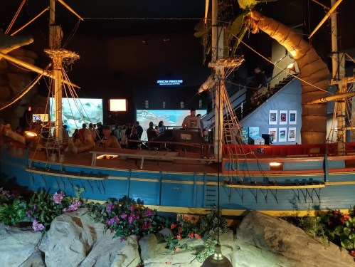 A large ship model displayed in a museum, surrounded by rocks and plants, with visitors in the background.