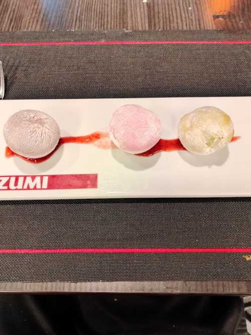 Three colorful mochi desserts on a white plate, drizzled with red sauce, set on a textured black tablecloth.