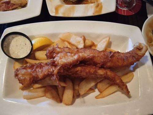 Two crispy fried fish fillets served on a plate with French fries, a lemon wedge, and a small cup of dipping sauce.
