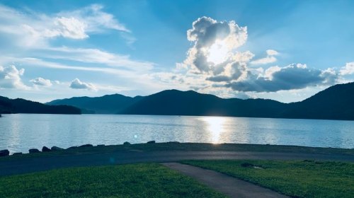 A serene lake scene with mountains in the background, under a bright sky with clouds and sunlight reflecting on the water.