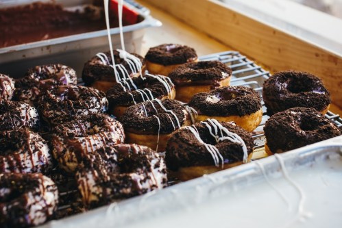 A tray of freshly made donuts topped with chocolate and crushed cookies, drizzled with white icing.