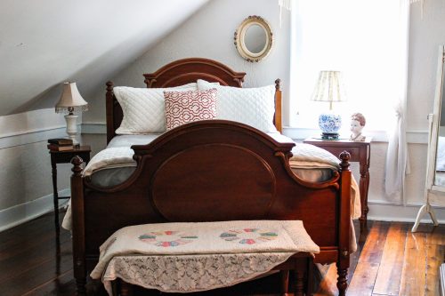 Cozy attic bedroom with a wooden bed, decorative pillows, a nightstand, and soft lighting from a window.
