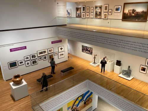 A spacious art gallery with various framed artworks, sculptures, and visitors exploring the exhibits on multiple levels.