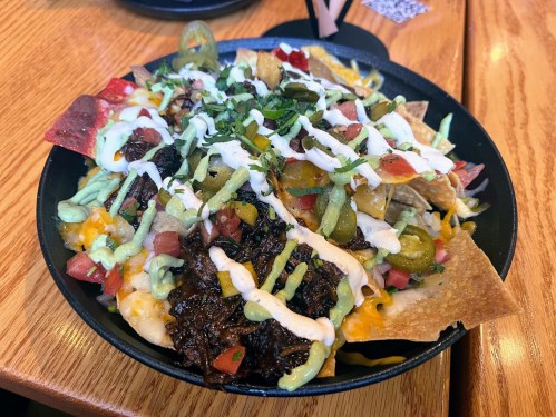 A close-up of a nacho dish topped with cheese, jalapeños, tomatoes, sour cream, and green sauce in a black bowl.