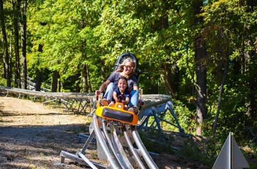 A woman and a child joyfully ride a roller coaster through a green, wooded area.