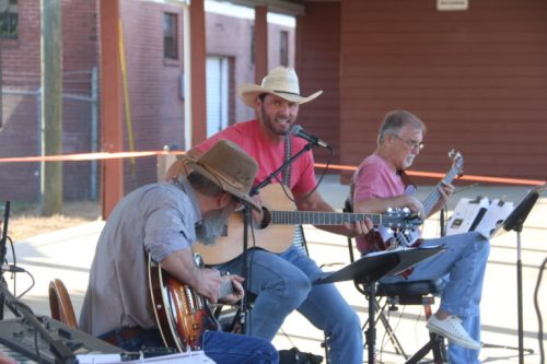 A trio of musicians performing outdoors, with one singing and playing guitar, while the others play instruments.