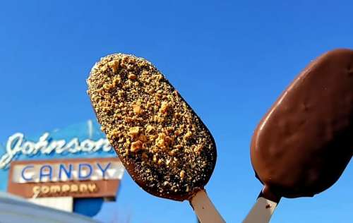 Two chocolate-covered ice cream bars with a candy shop sign in the background against a clear blue sky.
