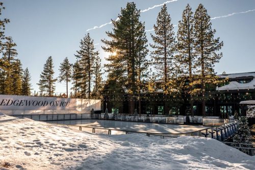 A snowy outdoor ice rink surrounded by tall pine trees and a building, with sunlight shining through the trees.